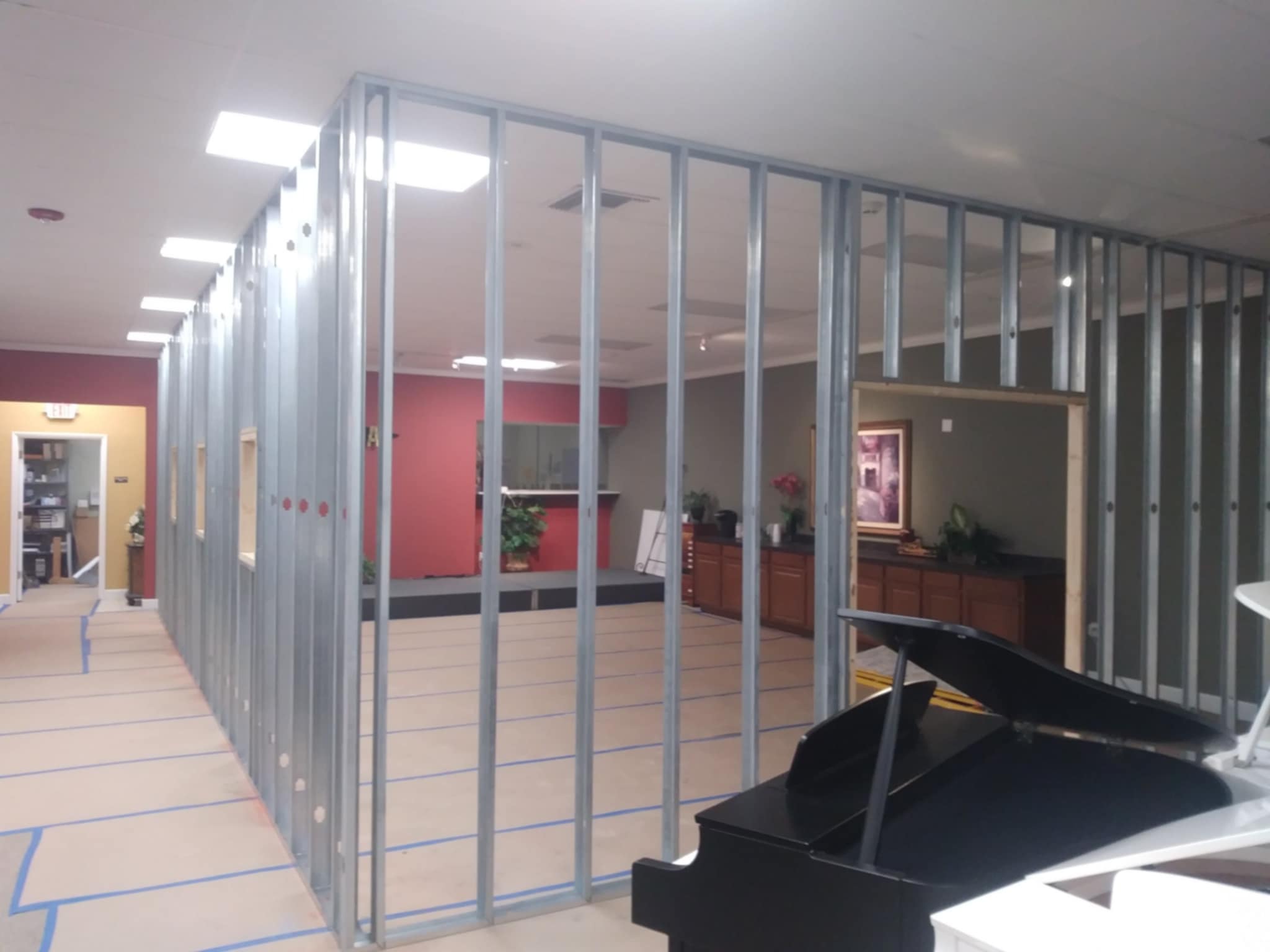 A framework of metal studs outlines a partially constructed wall in an office space, with a grand piano in the foreground. The area appears to be undergoing renovations.