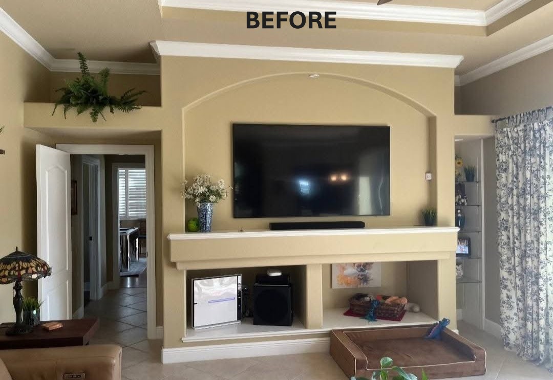 A living room with a large television mounted on a beige wall, featuring a decorative shelf below and a few plants. The space is well-lit, showcasing a cozy atmosphere.