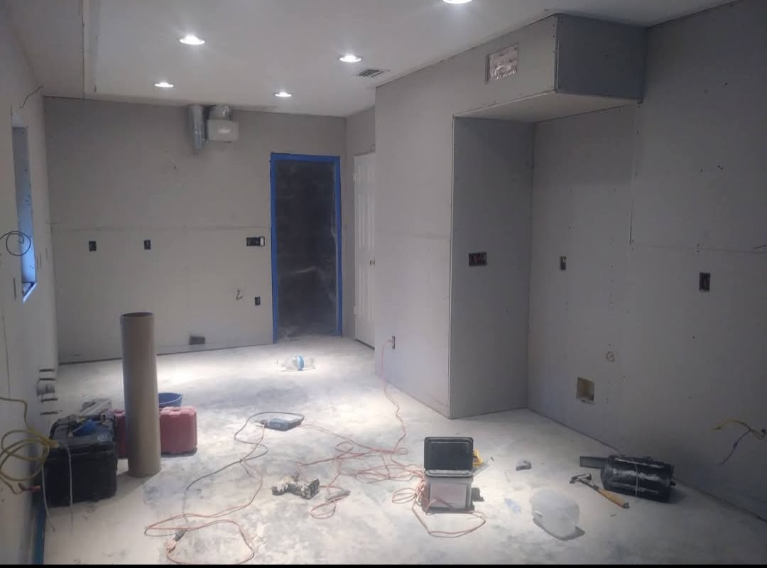 A partially renovated room with unfinished walls, scattered construction materials, and exposed wiring. The space is well-lit with ceiling lights.