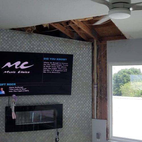 A partially renovated living room featuring a television mounted on a textured wall, a modern ceiling fan, and a fireplace. The room shows exposed framing, indicating ongoing construction or remodeling.