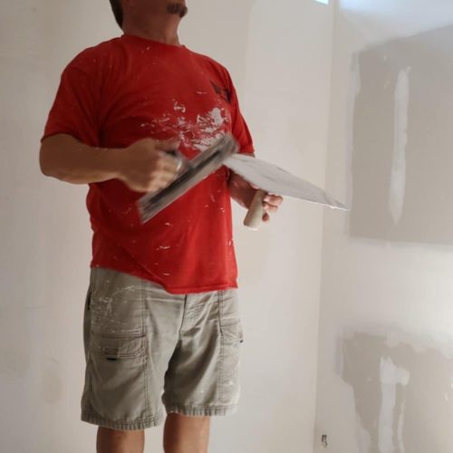 A worker applies finishing touches to drywall in a room with natural light coming through a window. He is standing on a platform and wearing a red shirt and shorts.