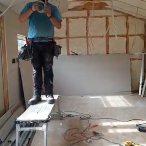 A person stands on a platform in a partially constructed room, holding a tool and preparing to work on the ceiling. Construction materials are scattered around the floor.