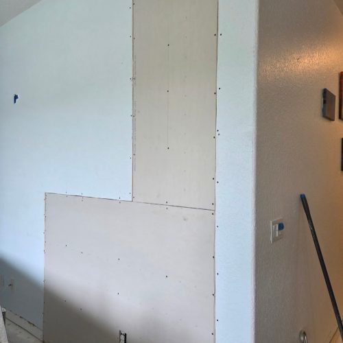 A section of drywall is installed on a wall, with part of the surface still unpainted. The area shows the transition between drywall and a painted wall.