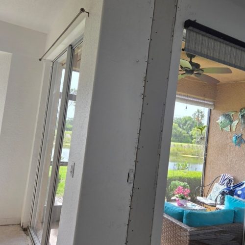 A vertical section of wall, partially unfinished, separates two areas in a bright interior space, with a view of a patio visible through sliding glass doors. The patio features a wicker chair and decorative items.