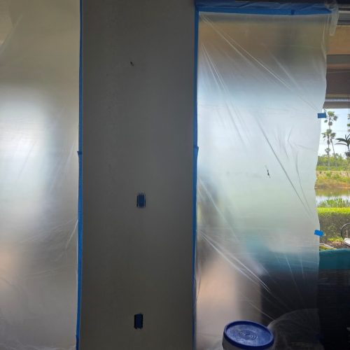 A wall partially covered with plastic sheeting, with a bucket of paint on the floor nearby. The setting appears to be a room undergoing renovation.