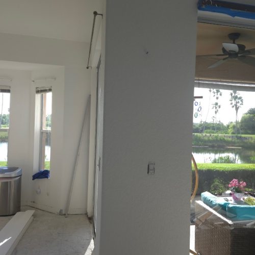 A renovated interior space featuring a newly finished wall, with natural light streaming in from large windows and a view of greenery outside. The area is clean and prepared for further decoration.