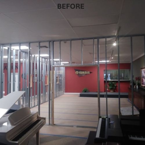 A room with a partially constructed interior, featuring exposed metal framing and a grand piano in the foreground. The wall in the background displays a Yamaha logo against a red backdrop.