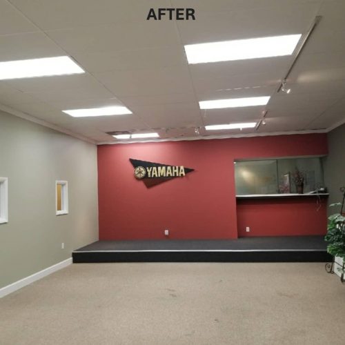 A renovated interior space featuring a red accent wall with a "Yamaha" logo, a small stage area, and new lighting. The room has a clean, modern look with carpeted flooring and a minimalist design.